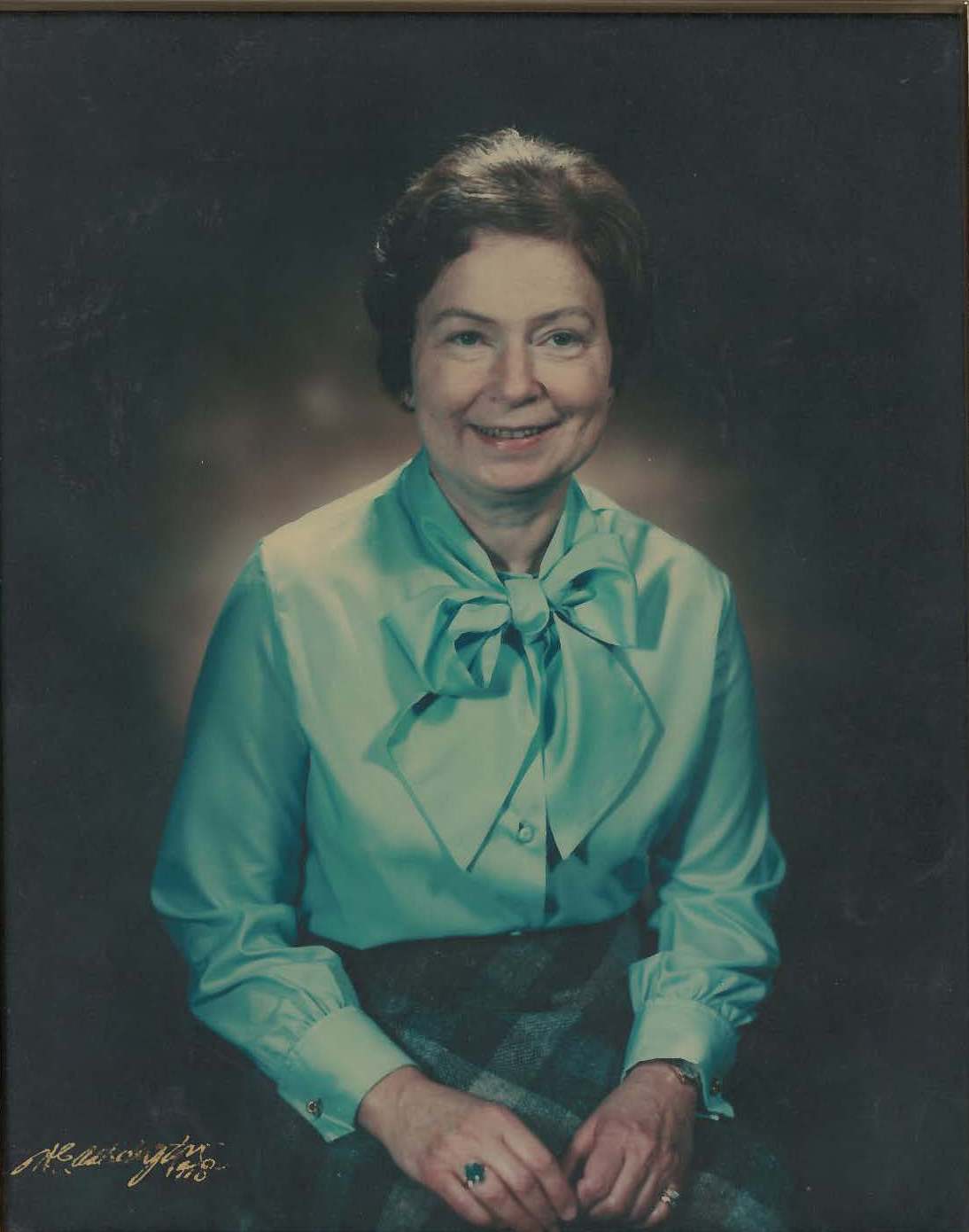 A smiling middle aged white woman with brown hair, wearing a light blue silk blouse with a bowtie and a tartan skirt and emerald ring set in gold